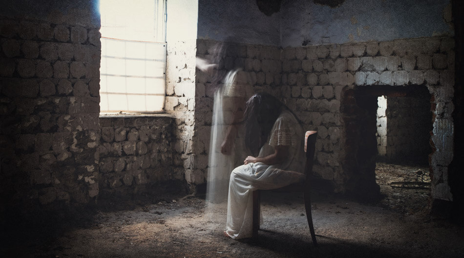 A person in a white dress is seated on a chair in a dilapidated, dimly lit room with stone walls, reminiscent of an old haunted asylum or hospital. Another translucent figure, possibly a ghost, stands by the window.
