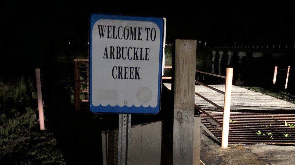 A nighttime view of a "Welcome to Arbuckle Creek" sign at the start of a old haunted looking bridge, illuminated by ambient light.