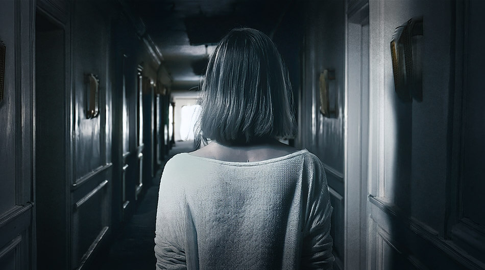 A female with short hair in a white sweater stands facing a dimly lit hallway, where the only light comes from a distant doorway, hinting the feeling of being watched which is a sign of a haunted house.