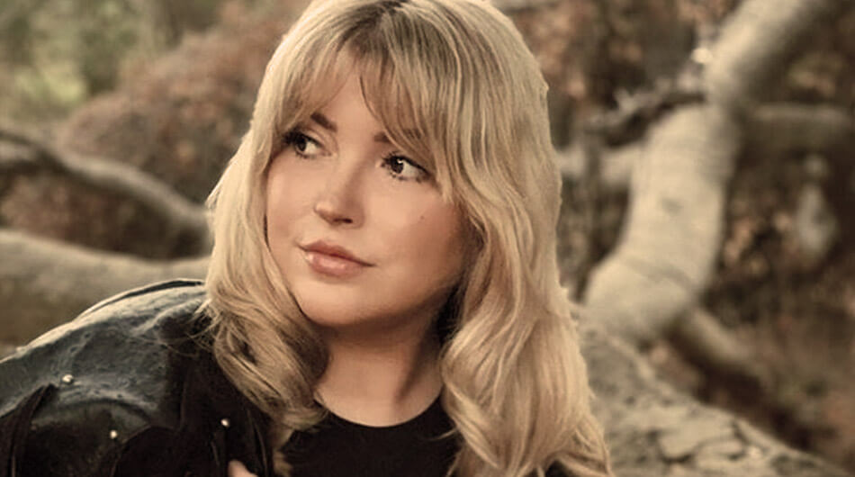 Jade Capasso wearing a dark top stands outdoors in a wooded area, looking to her left, as if she’s on the lookout for something mysterious—perhaps filming for her YouTube ghosthunters channel.