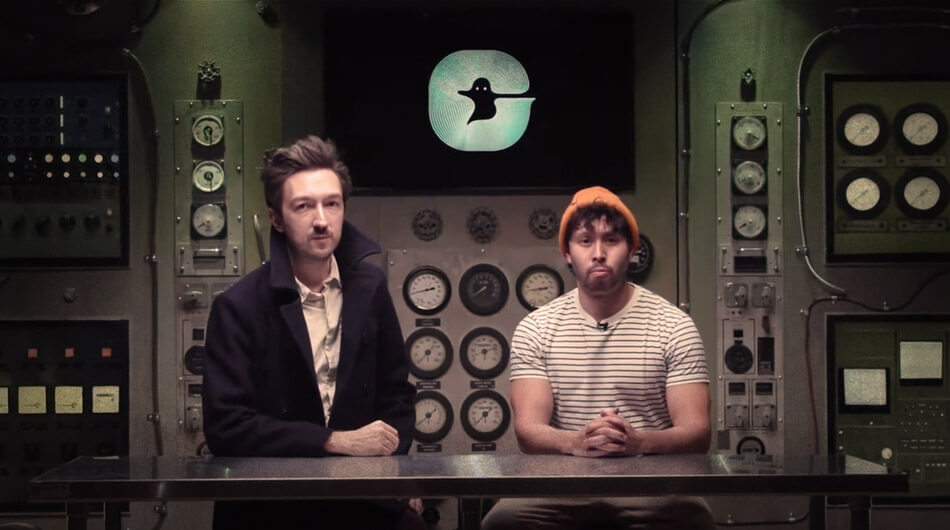 Two individuals sit at a desk in a dark room with various control panels and a screen showing a ghost icon in the background. One, wearing a dark jacket, is Shane Madej, while the other, sporting a striped shirt and orange beanie, is Ryan Bergara—arguably the best ghost hunters on YouTube.