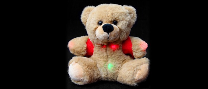 A plush BooBuddy Jr. Trigger Bear with a red vest sits against a black background, displaying small green and red lights on its surface. Perfect for any ghost hunting equipment list, this little bear is both adorable and intriguing.