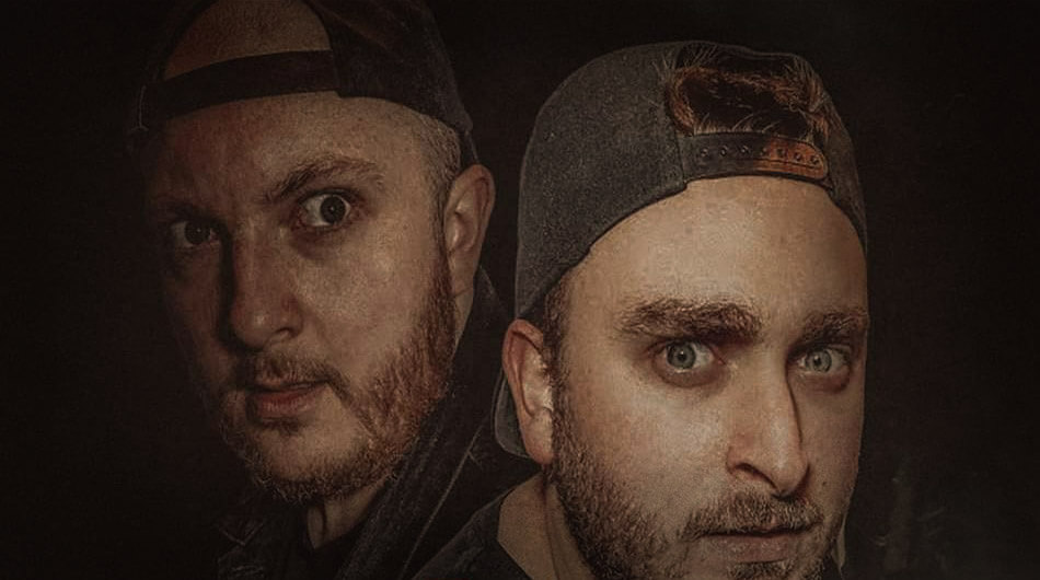Two men wearing backward baseball caps stare intensely. The man on the left has a beard and a shadowed face; the man on the right, identified as part of a popular YouTube ghost hunters channel called Ghost Theort, has a serious expression with bright lighting highlighting his face.