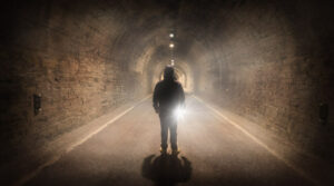 A person stands alone in a haunted tunnel, the dim light casting eerie shadows as they face forward, with a faint glow shining from behind.