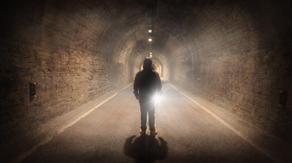 A person stands alone in a haunted tunnel, the dim light casting eerie shadows as they face forward, with a faint glow shining from behind.