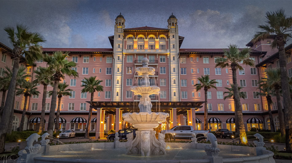 The Grand Hotel, the iconic Hotel Galvez, boasts an illuminated facade at dusk, with a multi-tiered fountain surrounded by palm trees—an enchanting scene that might make one wonder if it belongs among the haunted places in Houston.