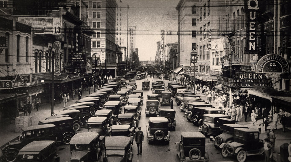 A busy 1920s urban Houston city with vintage cars, pedestrians, and tall buildings. Signs for "Queen" and "C.B. Daniels" are visible.