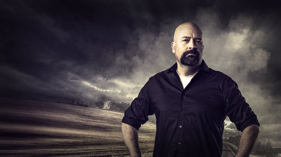 Jason Hawes standing outdoors in front of a dramatic cloudy sky and an open field. He is wearing a dark shirt and has a serious expression, with his hands on his hips. Now one the most popular and best ghost hunters on YouTube.