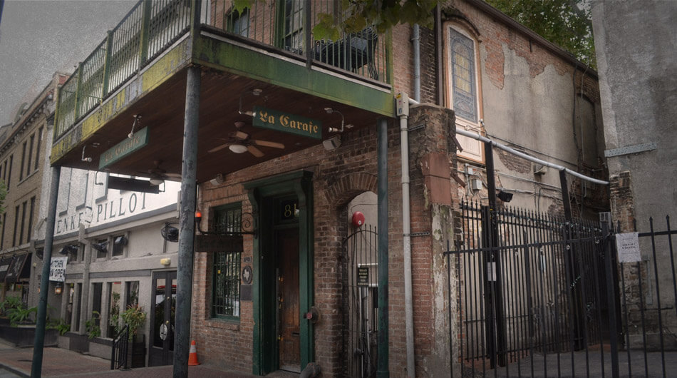 Street view of a two-story brick building with a balcony, green awning, and the iconic "La Carafe" sign, known for being one of the haunted places in Houston and oldest bar. Other buildings line the street, and a metal fence is visible to the right.