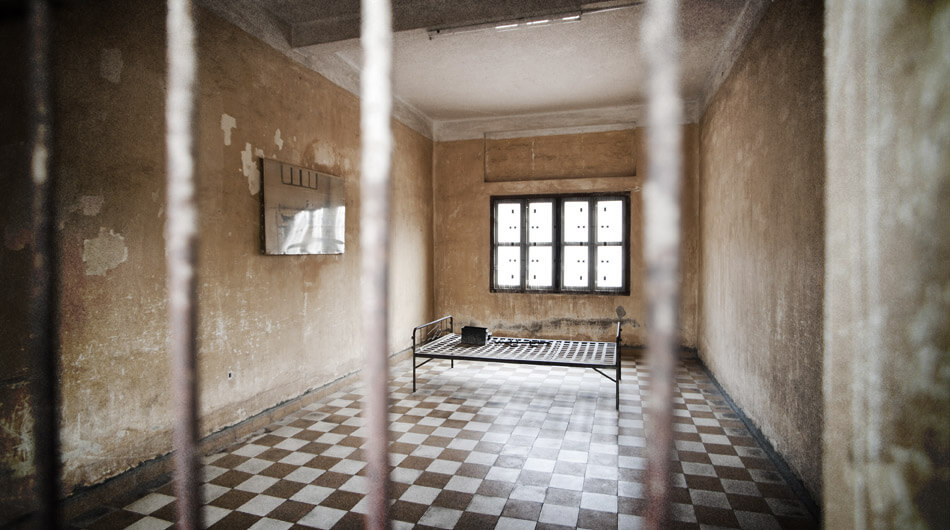 Possible a haunted jail with view through iron bars into a sparse room with a single metal-framed bed, checkered floor, peeling walls, and a barred window with a blurry light fixture.