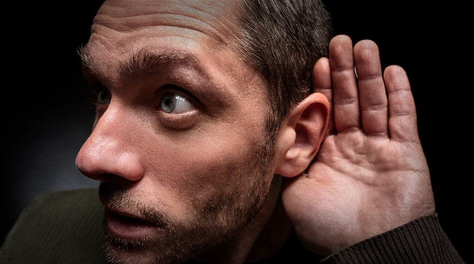 A man with short hair holds his hand to his ear, seemingly captivated by strange noises, against a dark background.