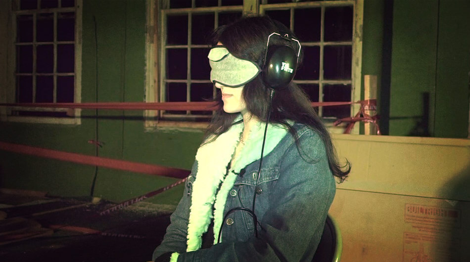 Person sitting indoors wearing a blindfold and headphones, using an Estes Method Kit, with windows and caution-taped area visible in the background.